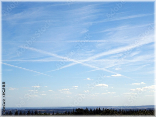 Vapor trails in a blue sky above the horizon. This is an immersive landscape with a line of  coniferous trees in the foreground shooted in 2019 in France mother country. Transparency white frame © Wijiji