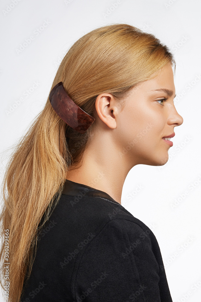 Cropped side view shot of blond girl with long hair, wearing black jacket. She pulling her hair with a dark brown rectangular hairpin with glossy overlay. The photo was taken on a white background. Cropped side view shot of blond girl with long hair, wearing black jacket. She pulling her hair with a dark brown rectangular hairpin with glossy overlay. The photo was taken on a white background.