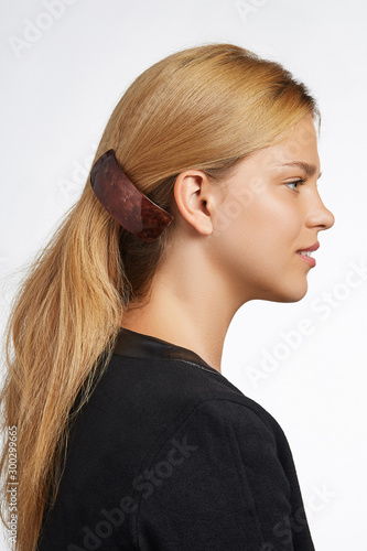 Cropped side view shot of blond girl with long hair, wearing black jacket. She pulling her hair with a dark brown rectangular hairpin with glossy overlay. The photo was taken on a white background. 