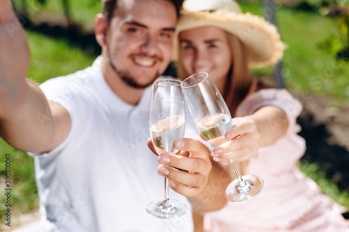 Beautiful couple is sitting on plaid holding glasses of wine and making selfies.