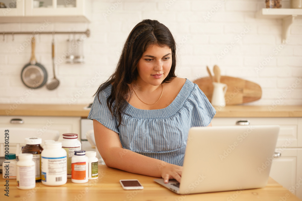 Attractive plus size young woman blogger working from home office, writing reviews online on new dietary supplements for body shape and weight loss on her account via social network, using laptop Attractive plus size young woman blogger working from home office, writing reviews online on new dietary supplements for body shape and weight loss on her account via social network, using laptop