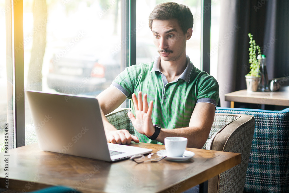 Stop it. Young serious businessman in green t-shirt sitting, working and looking at laptop screen and showing stop sign gesture. business and freelancing concept. indoor shot near window at daytime. Stop it. Young serious businessman in green t-shirt sitting, working and looking at laptop screen and showing stop sign gesture. business and freelancing concept. indoor shot near window at daytime.