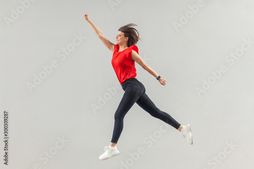 Full lenght of happy beautiful brunette young woman in casual red shirt jumping up in super hero pose and looking forward with toothy smile. indoor, studio shot, isolated on gray background.