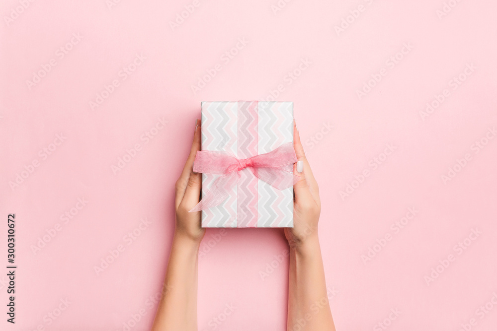 Female's hands holding striped gift box with colored ribbon on living coral background. Christmas concept or other holiday handmade present box, concept top view with copy space Female's hands holding striped gift box with colored ribbon on living coral background. Christmas concept or other holiday handmade present box, concept top view with copy space