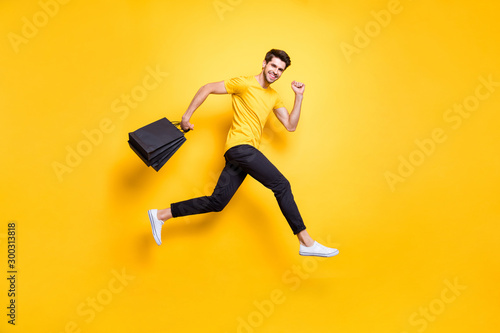 Full size photo of handsome guy jumping high holding many packages speed rushing sale shopping wear casual t-shirt black pants isolated yellow color background