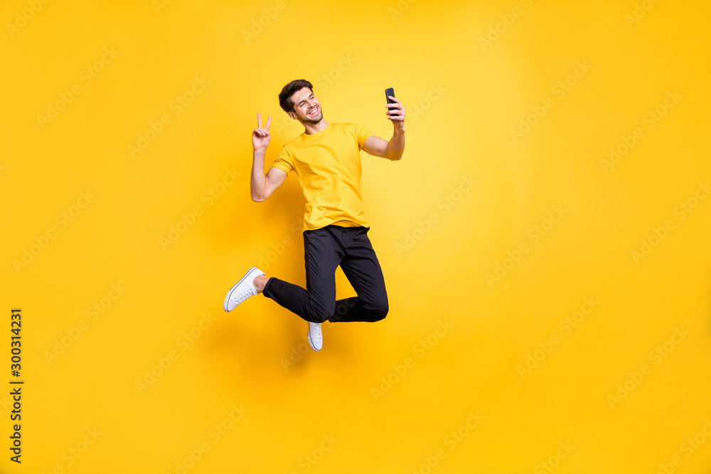 Full length photo of handsome guy jumping high holding telephone taking selfies showing v-sign symbol wear casual t-shirt pants isolated yellow color background