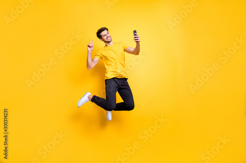 Full length photo of handsome guy jumping high holding telephone taking selfies showing v-sign symbol wear casual t-shirt pants isolated yellow color background photo