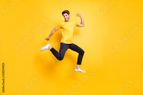 Full body photo of handsome guy jumping high waving arm old friends on streets glad to see them wear casual t-shirt black trousers isolated yellow color background