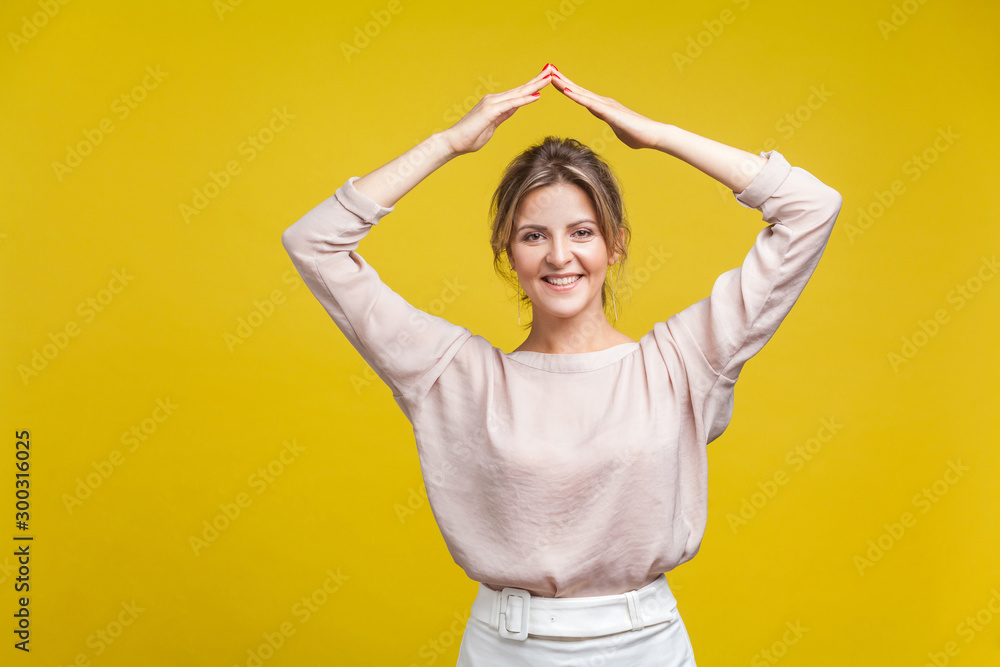 I’m in safety. Portrait of beautiful friendly woman with fair hair in casual blouse standing with arms over head, showing roof gesture, home symbol. indoor studio shot isolated on yellow background I’m in safety. Portrait of beautiful friendly woman with fair hair in casual blouse standing with arms over head, showing roof gesture, home symbol. indoor studio shot isolated on yellow background