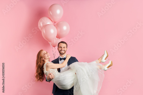 Attractive redhaired woman wearing white wedding dress sit on man's hands. Cheerful smiling couple happy together, marriage proposal