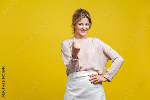 Come here! Portrait of playful happy young woman with fair hair in casual beige blouse standing, calling with finger gesture, inviting to come in. indoor studio shot isolated on yellow background