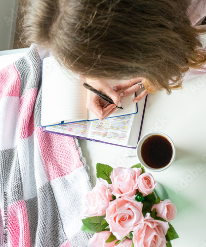 Woman doing devotional Bible study and writing in a journal. Pink roses and coffe on a white table. 