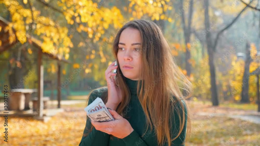 Young attractive brunette woman talking on phone and holding pack of money. Girl enjoys lot of cash. Huge wealth of money. Employee salary. Cash money payment. Successful business concept. 4K footage Young attractive brunette woman talking on phone and holding pack of money. Girl enjoys lot of cash. Huge wealth of money. Employee salary. Cash money payment. Successful business concept. 4K footage
