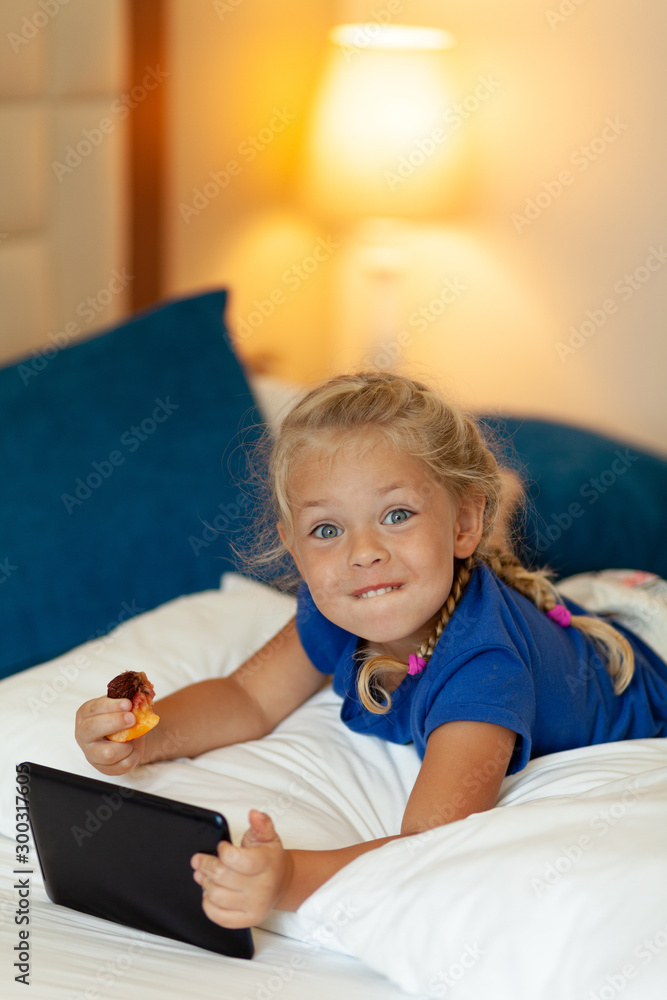 Little pretty girl holding tablet in her hands eats fruit and lies on bed (sofa) at home. Mobile game, entertainment and communication via Internet. Little pretty girl holding tablet in her hands eats fruit and lies on bed (sofa) at home. Mobile game, entertainment and communication via Internet.