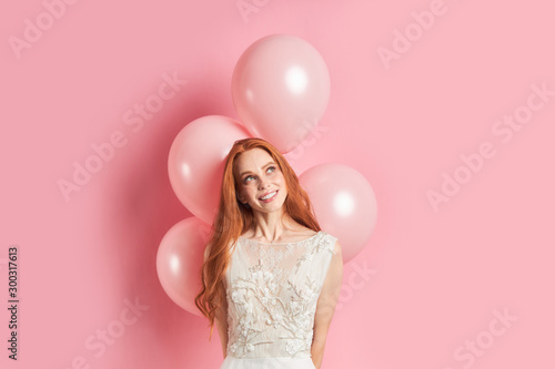Young and beautiful woman look up isolated over pink background. Air balloons behind. Dreamy female with long red hair.