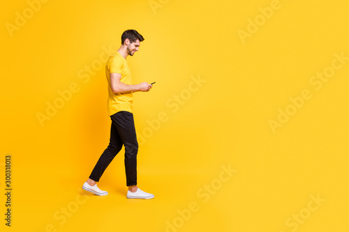 Full length body size photo of cheerful attractive man with bristle smiling toothily focused on reading feednews on his phone wearing yellow t-shirt white footwear isolated bright color background