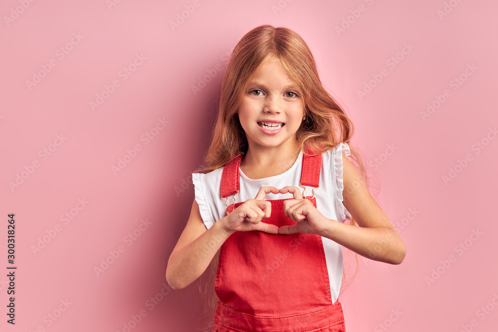 Portrait of attractive girl wearing red dress, having long hair stand showing heart isolated over pink background. Portrait of attractive girl wearing red dress, having long hair stand showing heart isolated over pink background.