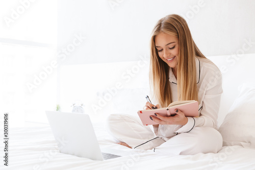 Photo of blonde smiling woman make notes and using laptop