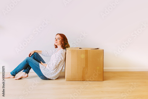 Young attractive girl moving into the new apartment. Sitting on the floor with big carton box in an empty room on a white wall background.