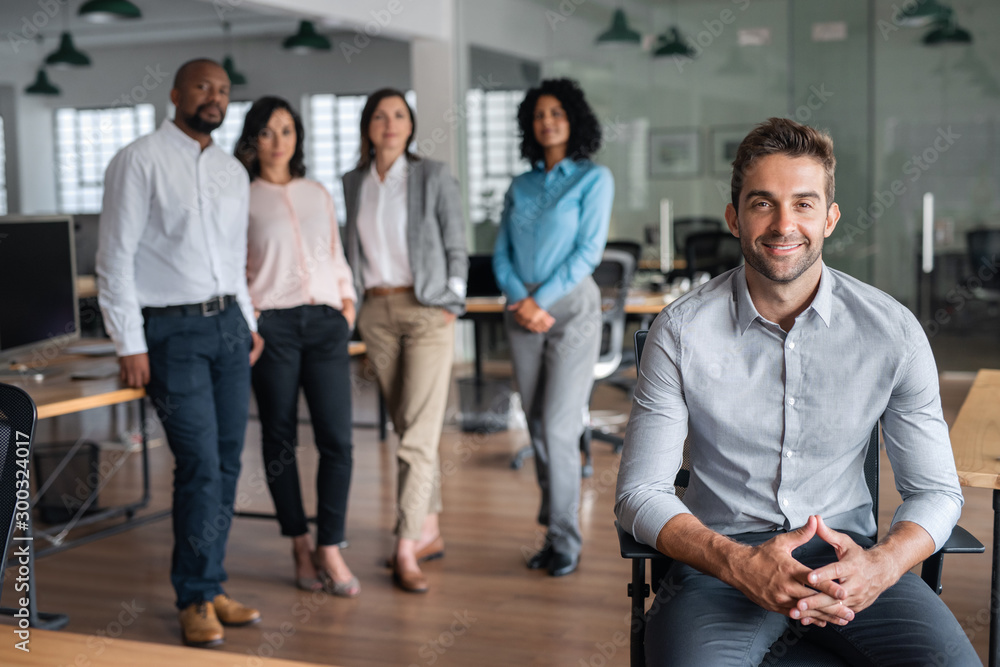 Smiling businessman sitting in an office with coworkers behind him Smiling businessman sitting in an office with coworkers behind him