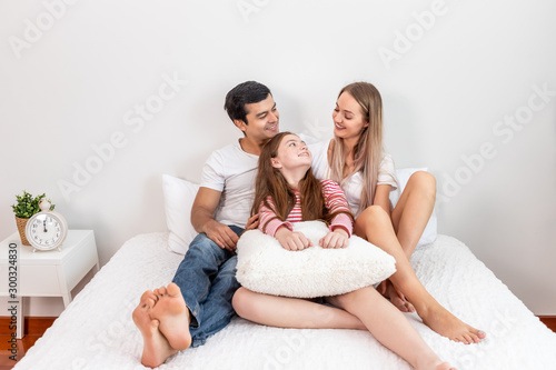 Happy family, including father mother and daughter, are sitting on bed with smile, looking at each other