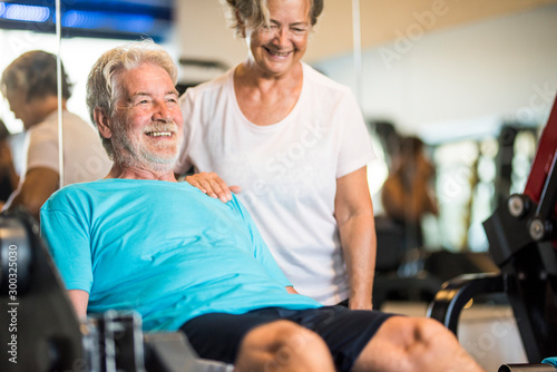 woman helping his man at the gym to do exercise with his legs - active couple of pensioners doing exercises together - healthy lifestyle
