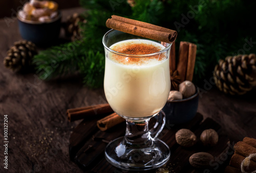 New Year or Christmas Eggnog cocktail - hot winter or autumn drink with milk, eggs and dark rum, sprinkled with cinnamon and nutmeg in a glass on wooden background, festive decoration