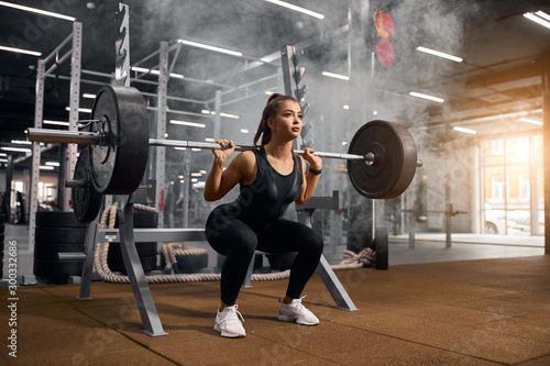 Fototapeta Naklejka Na Ścianę i Meble -  Beautiful young powerlifter squatting in modern fitness studio using heavy barbell, looking aside expressing self assurance, professional sport concept, white smoke in the air