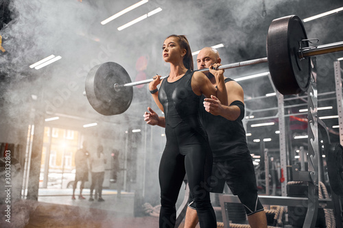 Low angle shot of sporty friends training in underground garage, standing in white smoke, holding heavy black barbell, brutal professional sportsman supporting young female partner, indoor shot photo