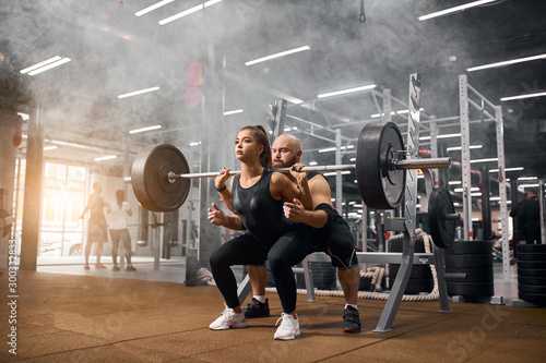 Brutal male weightlifter standing behind young sporty woman backing up squatting process with heavy barbell in brightly lighted gym, practicing in white smoke, shot from below, indoor shot