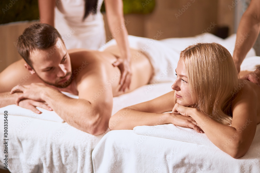Lovely young couple having pleasure time in beauty treatment spa salon, lying under soft fresh towels, looking at each other, receiving aromatherapy massage, side indoor shot Lovely young couple having pleasure time in beauty treatment spa salon, lying under soft fresh towels, looking at each other, receiving aromatherapy massage, side indoor shot