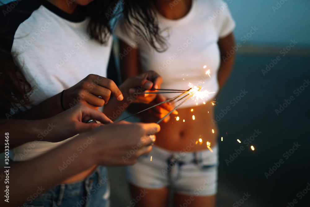 Young happy girls stands on beach with sparkler in sunset light. Teenage girls with sparklers celebrate and laugh. Party, holidays, nightlife and people concept.