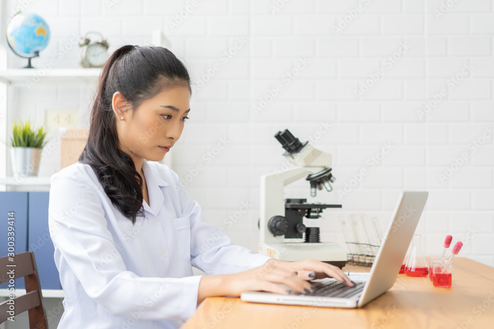 Asian female doctor working on laptop with a microscope for an exam at hospital giving patient convenience online service advice, smiling write a prescription , healthcare, preventing disease concept Asian female doctor working on laptop with a microscope for an exam at hospital giving patient convenience online service advice, smiling write a prescription , healthcare, preventing disease concept