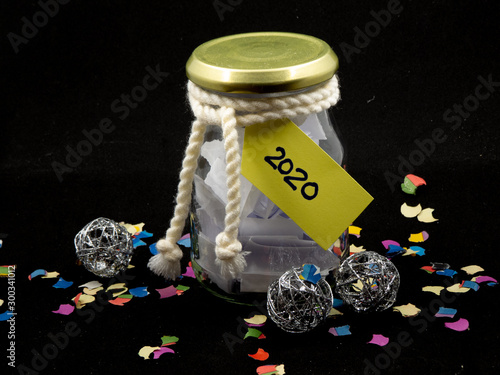 Glass jar full of paper shreds and tag reading 2020 surrounded by colorful confetti and silver ornaments isolated on black background
