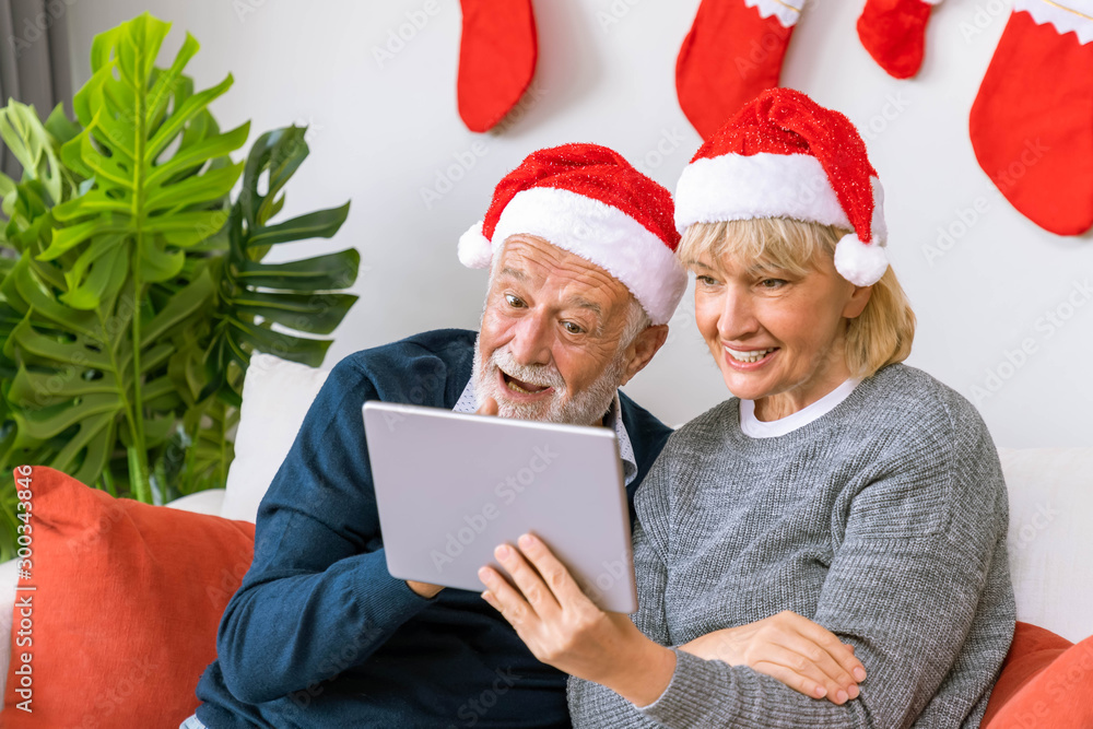 Senior couple using tablet to video phone call to greeting their family for Christmas festival, sitting on sofa with decoration and tree Senior couple using tablet to video phone call to greeting their family for Christmas festival, sitting on sofa with decoration and tree