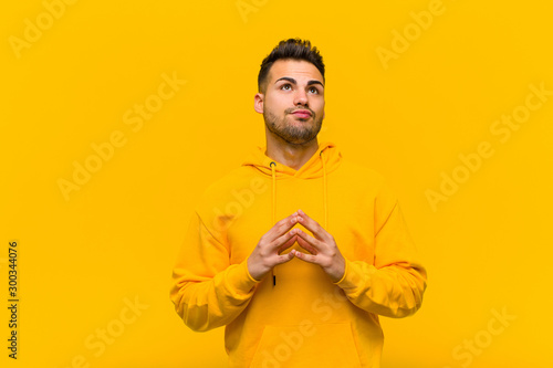 young hispanic man scheming and conspiring, thinking devious tricks and cheats, cunning and betraying against orange wall