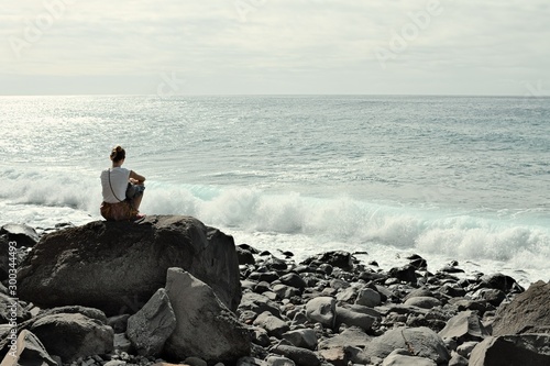 Isolated young woman thinking on a rock in the shoreline of the Atlantic Ocean coast (Madeira, Portugal, Europe)