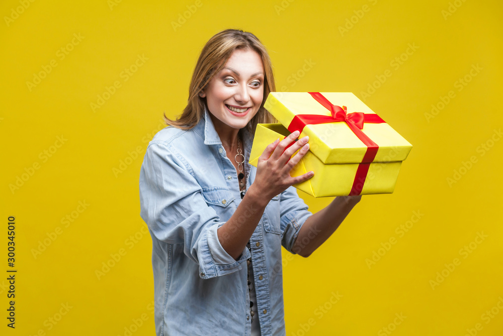 Long awaited present. Portrait of curious woman in denim shirt looking inside gift box, checking what's inside, unboxing with satisfied happy expression. studio shot isolated on yellow background Long awaited present. Portrait of curious woman in denim shirt looking inside gift box, checking what's inside, unboxing with satisfied happy expression. studio shot isolated on yellow background