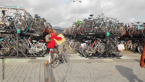 AMSTERDAM, Netherlands - October 2019: young woman on a bicycle rides up to a large bicycle park. A black man walks past.