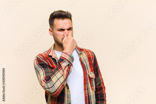 young hispanic man feeling disgusted, holding nose to avoid smelling a foul and unpleasant stench against isolated background