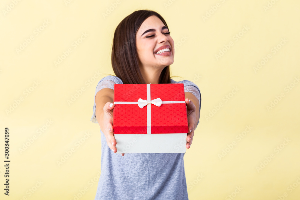 Fototapeta premium Happy brunette woman in casual holding gift box and looking at the camera while enjoys over yellow background. Young woman hold gift in christmas style