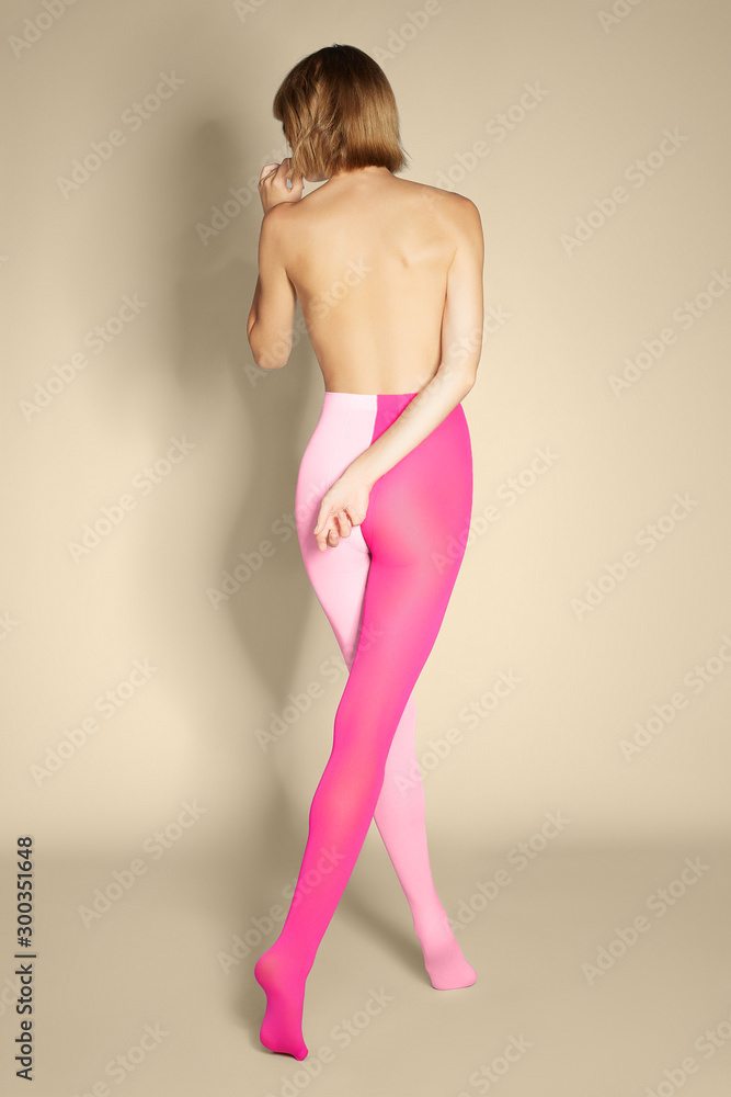 Back view of a blonde lady in pink two-tone tights. The slim shirtless woman with a short hairdo is posing on tiptoes on the beige background. 