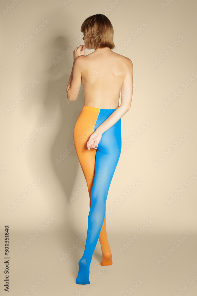 Back view of a blonde lady in blue and orange tights. The slim shirtless woman with a short hairdo is posing on tiptoes on the beige background. Back view of a blonde lady in blue and orange tights. The slim shirtless woman with a short hairdo is posing on tiptoes on the beige background.