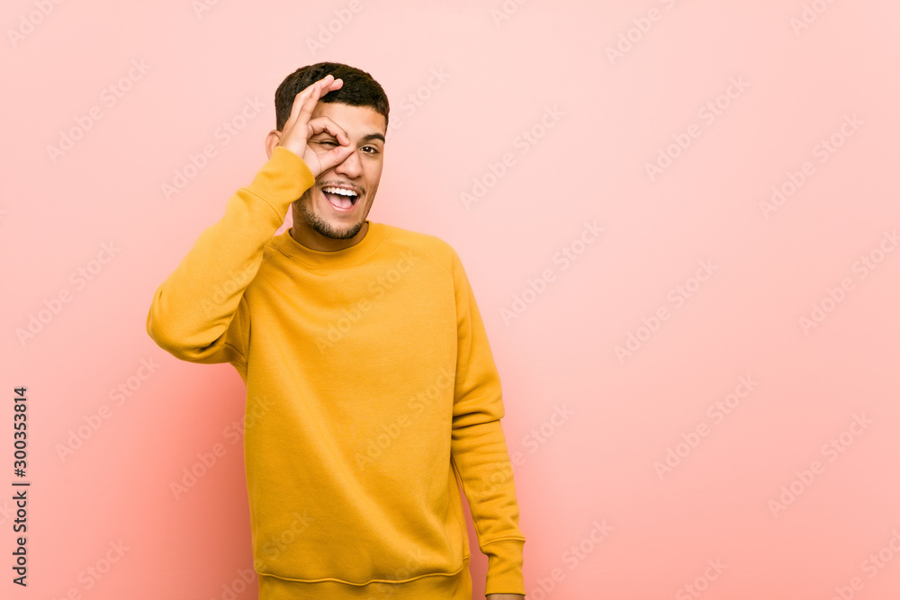 Young hispanic man excited keeping ok gesture on eye. Young hispanic man excited keeping ok gesture on eye.