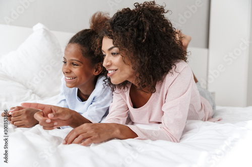 Image of african american woman and her little daughter lying in bed at home