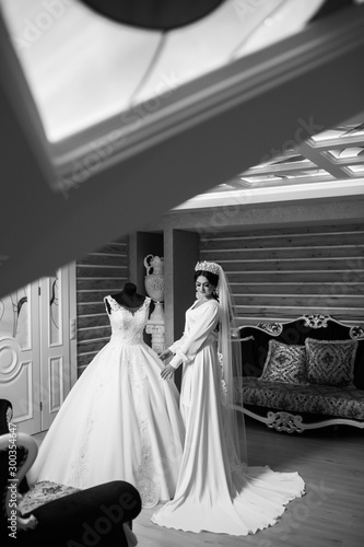 Black and white photo. Luxury bride while preparing morning. Bride in white robe standing near the mannequin with wedding dress in the room in the morning. Bride with make up and hair style with crown