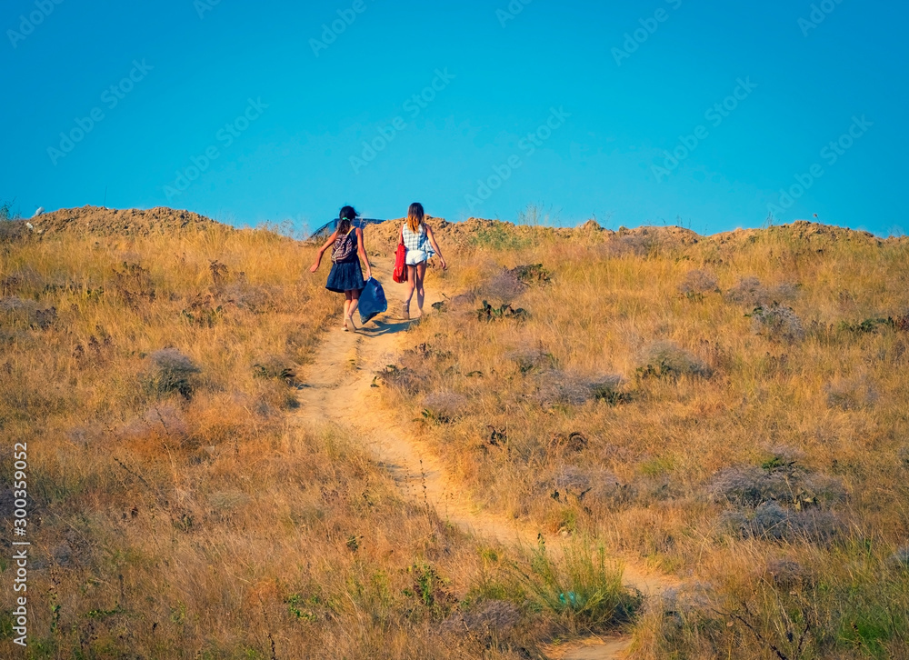 Two girls with heavy bags climb to the top of the hill. People walking from the beach to the car park on a hot summer day on the sea coast. Overcoming difficulties concept.