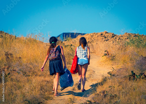 Two girls with heavy bags climb to the top of the hill. People walking from the beach to the car park on a hot summer day on the sea coast. Overcoming difficulties concept.
