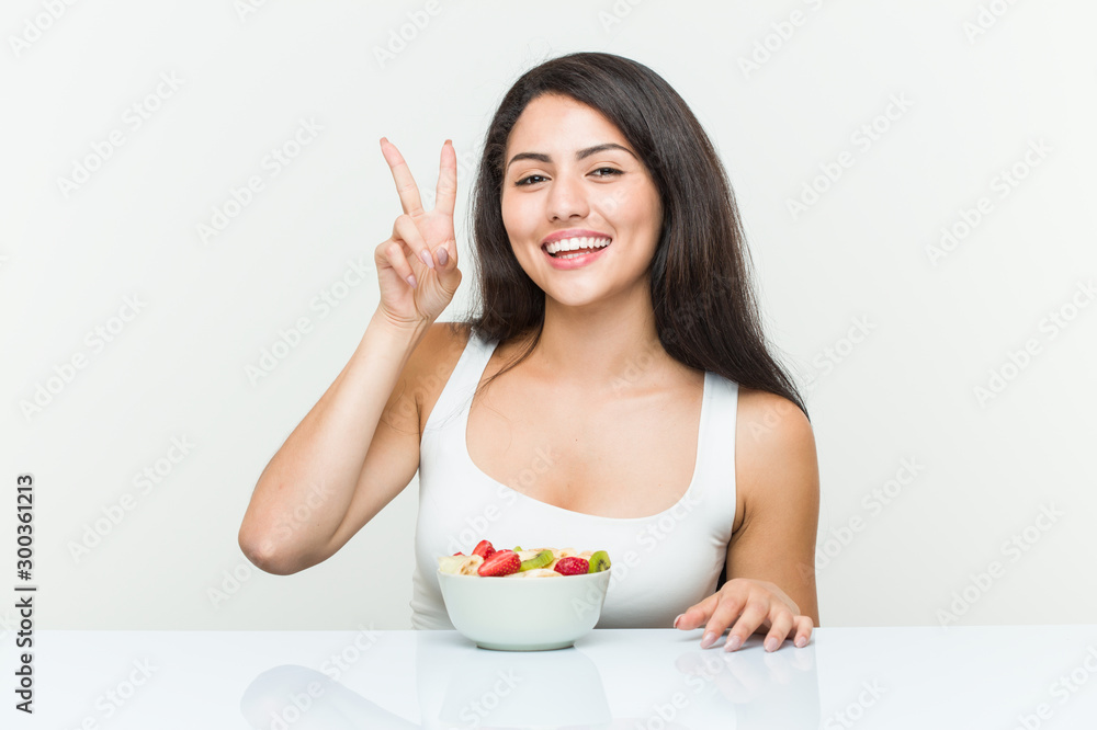 Young hispanic woman eating a fruit bowl showing victory sign and smiling broadly. Young hispanic woman eating a fruit bowl showing victory sign and smiling broadly.