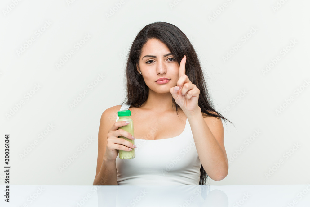 Young hispanic woman holding a vegetable drink showing number one with finger. Young hispanic woman holding a vegetable drink showing number one with finger.